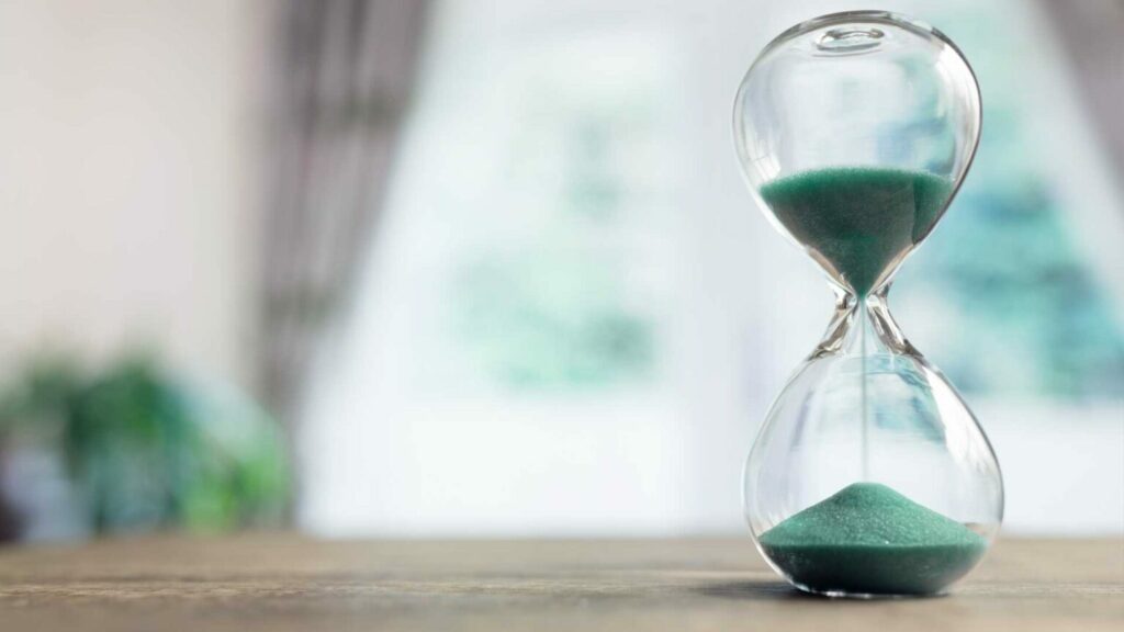 An hourglass with green sand placed on a wooden surface, symbolizing the passage of time. The blurred background gives a soft and minimalistic aesthetic, representing concepts of urgency, time management, and change.