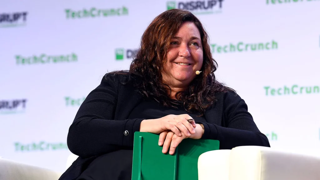 A woman speaking at a TechCrunch Disrupt event, holding a green notebook and smiling image